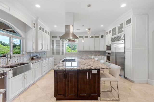 kitchen with hanging light fixtures, built in appliances, a kitchen island, light stone counters, and sink