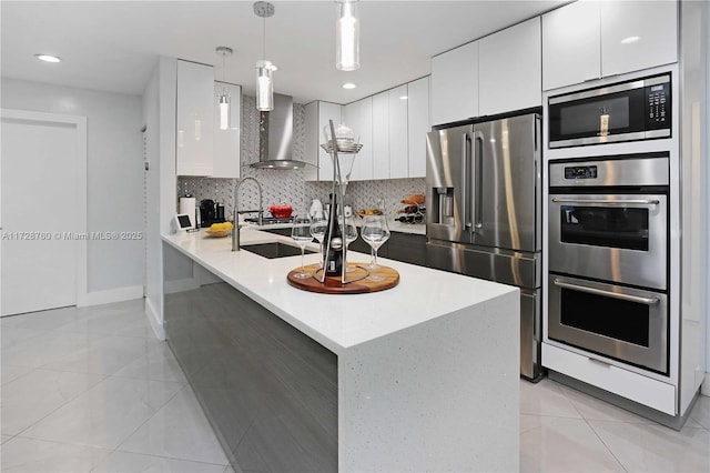 kitchen with white cabinetry, hanging light fixtures, appliances with stainless steel finishes, wall chimney range hood, and backsplash