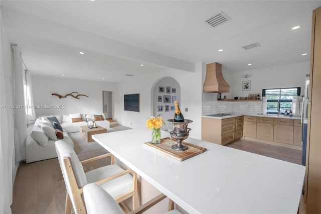 kitchen featuring custom exhaust hood, tasteful backsplash, light wood-type flooring, stainless steel gas cooktop, and light brown cabinetry