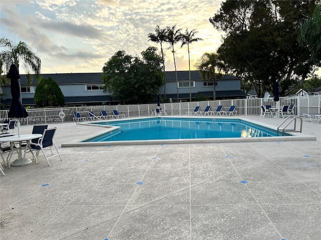 pool at dusk with a patio area, fence, and a community pool