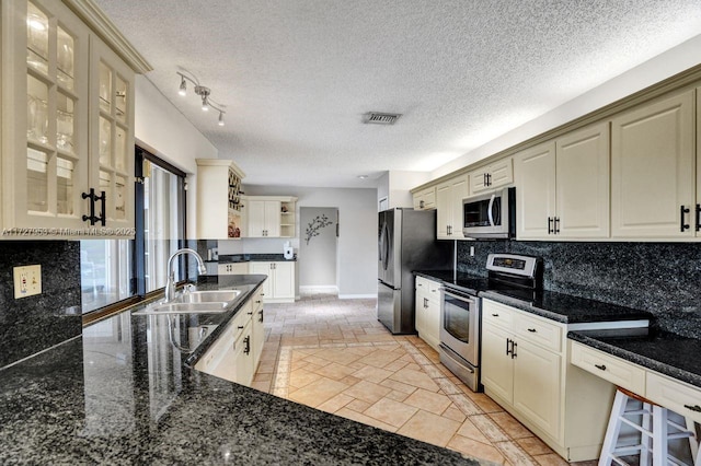 kitchen with stainless steel appliances, dark stone counters, sink, backsplash, and cream cabinetry