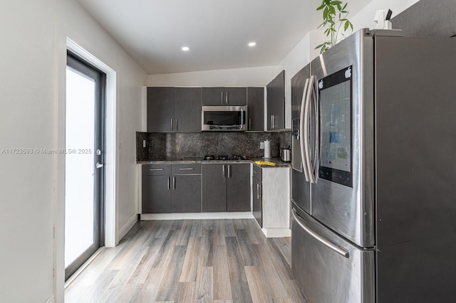 kitchen with appliances with stainless steel finishes, decorative backsplash, light wood-type flooring, dark stone counters, and vaulted ceiling