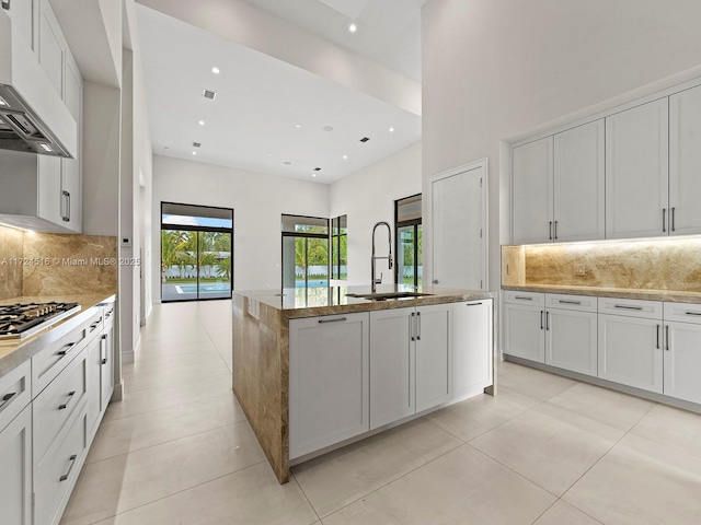 kitchen featuring range hood, decorative backsplash, sink, an island with sink, and white cabinets