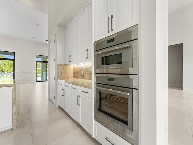 kitchen with light stone countertops, stainless steel double oven, white cabinetry, tasteful backsplash, and light tile patterned flooring