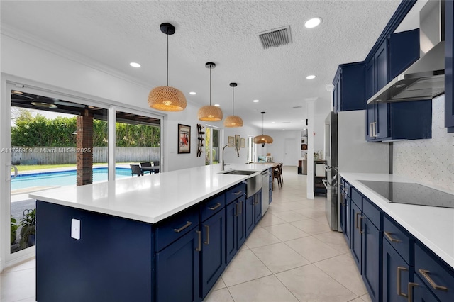 kitchen with pendant lighting, wall chimney range hood, a center island with sink, and blue cabinets