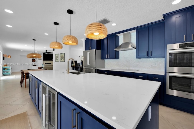 kitchen featuring an island with sink, appliances with stainless steel finishes, blue cabinetry, wall chimney range hood, and pendant lighting