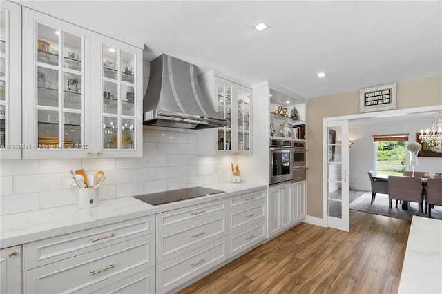 kitchen with black electric cooktop, stainless steel oven, white cabinets, and wall chimney range hood