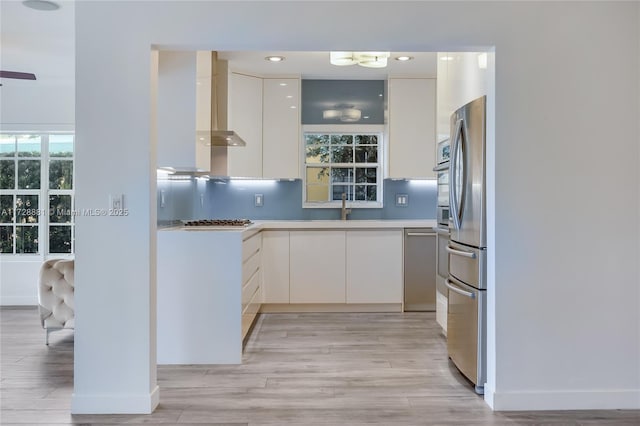 kitchen featuring sink, white cabinetry, light wood-type flooring, appliances with stainless steel finishes, and wall chimney range hood