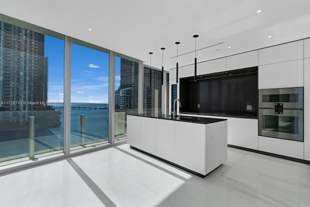 kitchen with a center island with sink, a wall of windows, decorative light fixtures, double oven, and white cabinets