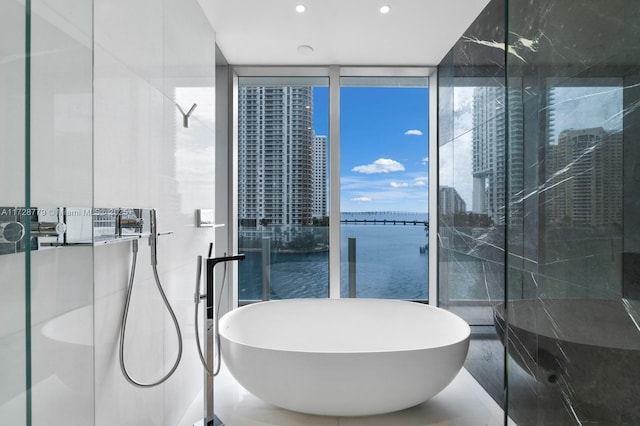 bathroom with a wall of windows, a water view, a tub, and tile walls