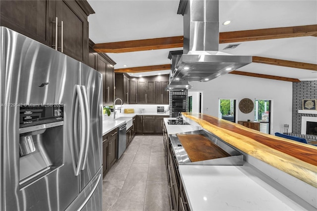 kitchen with appliances with stainless steel finishes, backsplash, island range hood, lofted ceiling with beams, and a brick fireplace