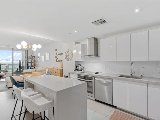 kitchen with appliances with stainless steel finishes, hanging light fixtures, wall chimney range hood, white cabinets, and sink