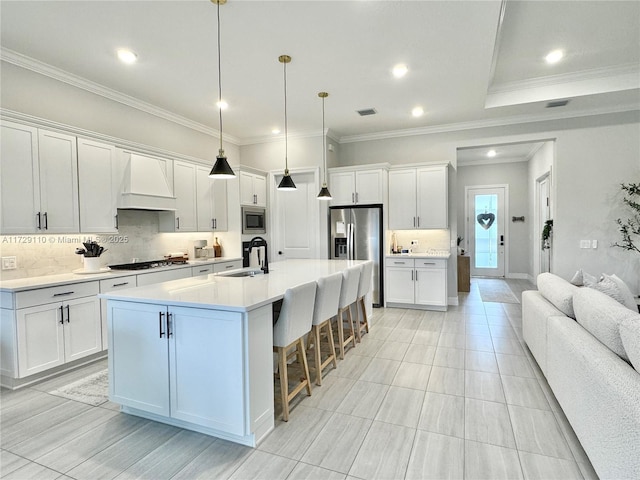 kitchen featuring appliances with stainless steel finishes, premium range hood, white cabinets, sink, and an island with sink