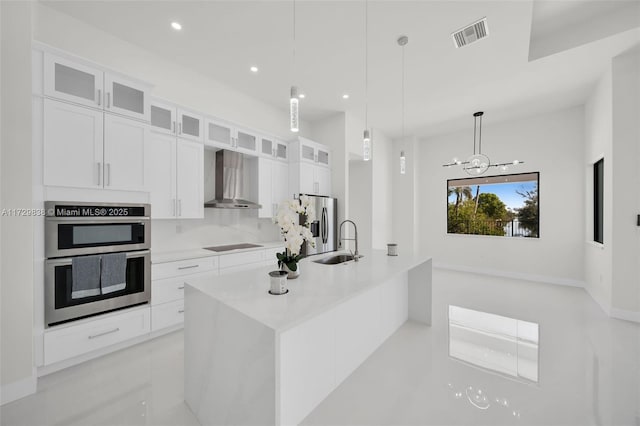 kitchen featuring hanging light fixtures, white cabinets, wall chimney range hood, stainless steel appliances, and a center island with sink