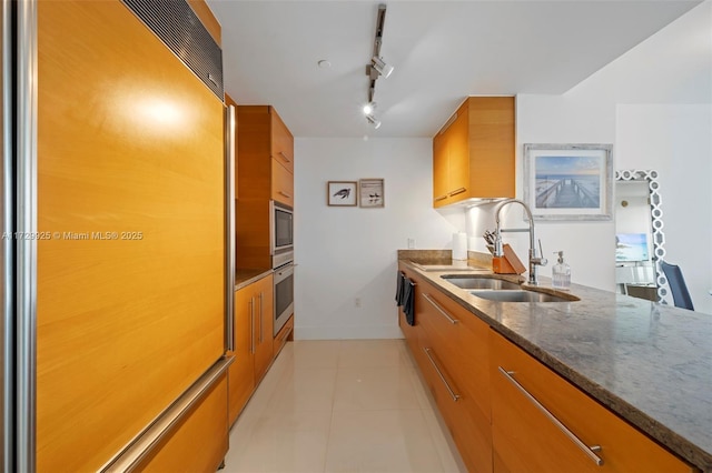 kitchen featuring stainless steel oven, rail lighting, sink, paneled built in refrigerator, and dark stone counters