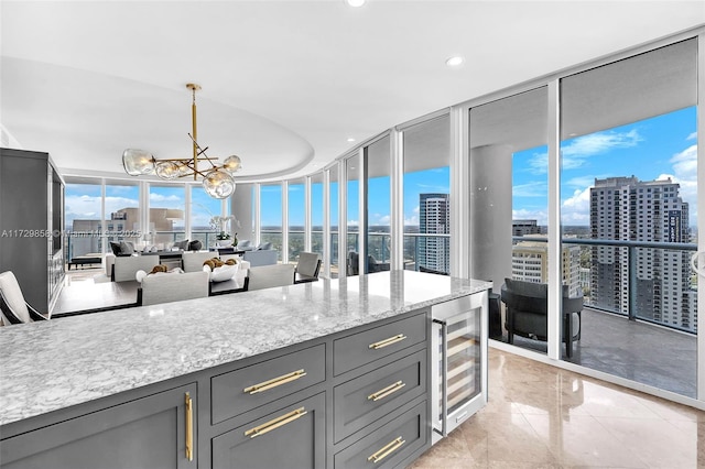 kitchen featuring wine cooler, an inviting chandelier, pendant lighting, a wall of windows, and light stone countertops