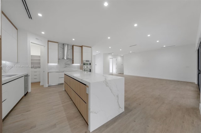 kitchen featuring wall chimney exhaust hood, white cabinets, a spacious island, and light stone counters