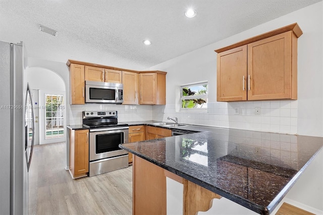 kitchen with light hardwood / wood-style flooring, appliances with stainless steel finishes, decorative backsplash, kitchen peninsula, and dark stone counters