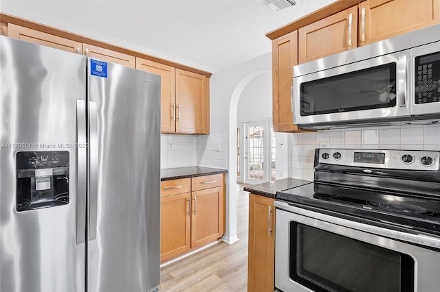 kitchen with dark stone countertops, stainless steel appliances, decorative backsplash, light wood-type flooring, and light brown cabinets