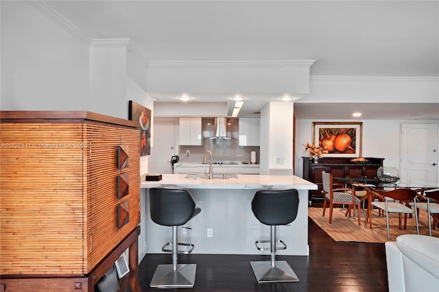 kitchen featuring dark wood-type flooring, a kitchen bar, sink, white cabinetry, and wall chimney range hood