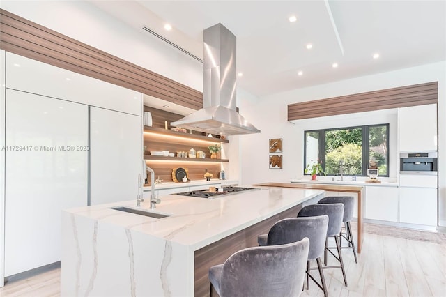 kitchen featuring stainless steel gas stovetop, white cabinets, island exhaust hood, light stone counters, and a kitchen island with sink