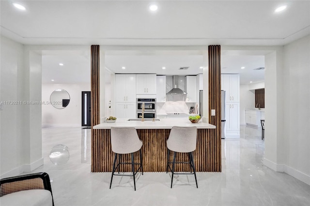 kitchen featuring a breakfast bar area, fridge, stainless steel double oven, wall chimney range hood, and white cabinets