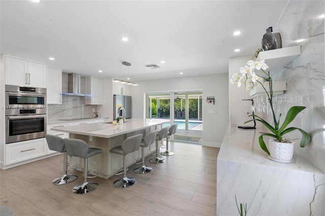 kitchen with decorative light fixtures, white cabinetry, an island with sink, stainless steel appliances, and wall chimney range hood