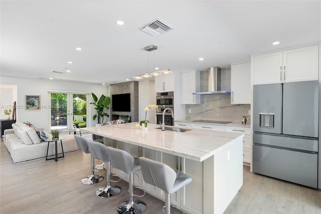 kitchen with white cabinetry, wall chimney range hood, light stone counters, stainless steel appliances, and a center island with sink