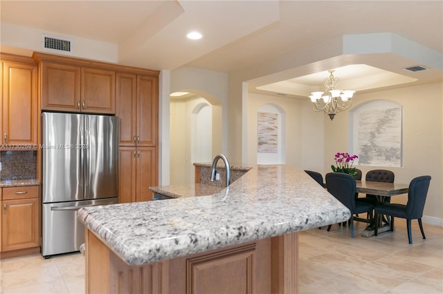 kitchen featuring light stone countertops, tasteful backsplash, an island with sink, stainless steel fridge, and a tray ceiling