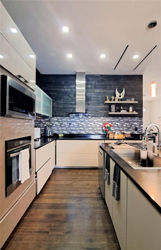 kitchen with pendant lighting, appliances with stainless steel finishes, wall chimney exhaust hood, dark wood-type flooring, and white cabinetry