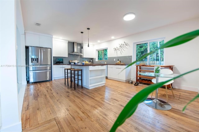 kitchen featuring stainless steel refrigerator with ice dispenser, wall chimney range hood, white cabinets, pendant lighting, and a healthy amount of sunlight