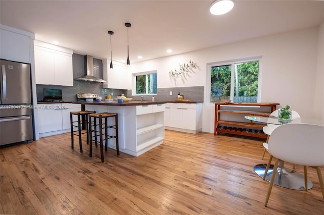 kitchen with appliances with stainless steel finishes, wall chimney exhaust hood, decorative light fixtures, white cabinetry, and light hardwood / wood-style flooring