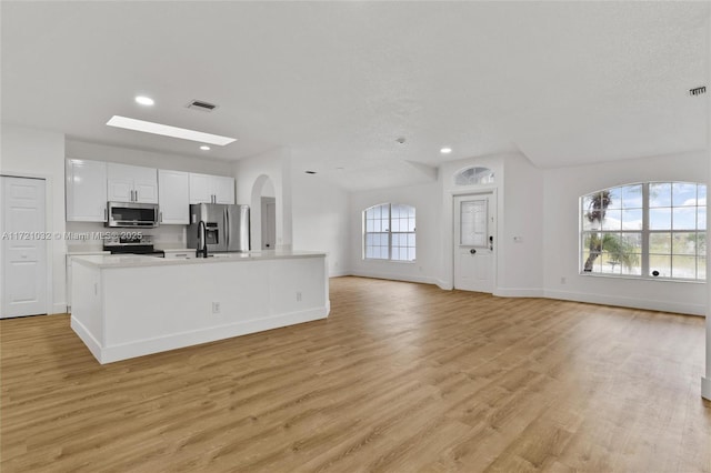 kitchen with appliances with stainless steel finishes, a skylight, white cabinets, light wood-type flooring, and an island with sink