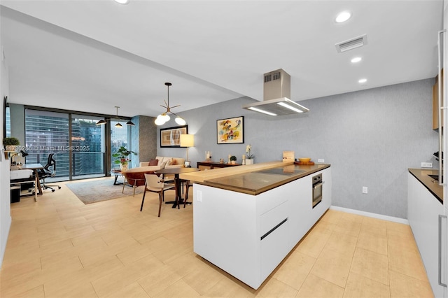 kitchen with pendant lighting, a wall of windows, white cabinetry, island range hood, and stainless steel oven