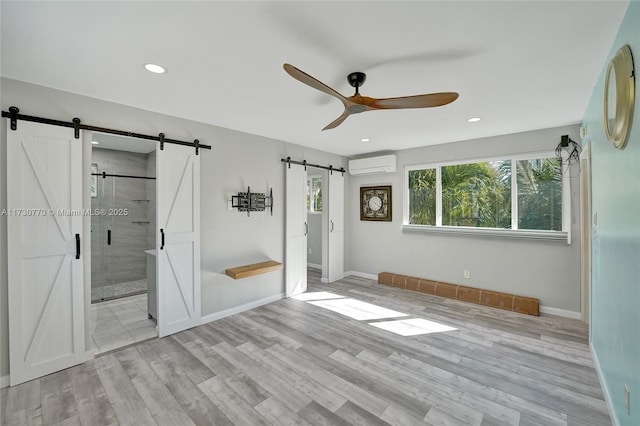 unfurnished bedroom featuring ensuite bathroom, an AC wall unit, ceiling fan, a barn door, and light hardwood / wood-style floors