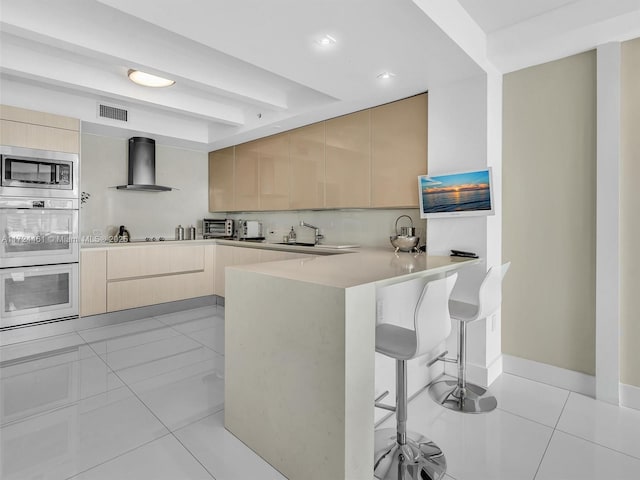 kitchen featuring wall chimney range hood, stainless steel microwave, kitchen peninsula, and light brown cabinets