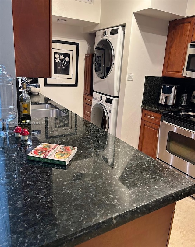 kitchen with appliances with stainless steel finishes, sink, light tile patterned floors, dark stone counters, and stacked washing maching and dryer