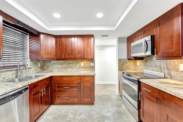 kitchen featuring sink, light stone counters, appliances with stainless steel finishes, a tray ceiling, and decorative backsplash