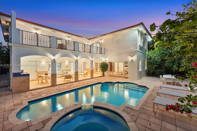 back house at dusk featuring a balcony, a pool with hot tub, a patio area, and french doors