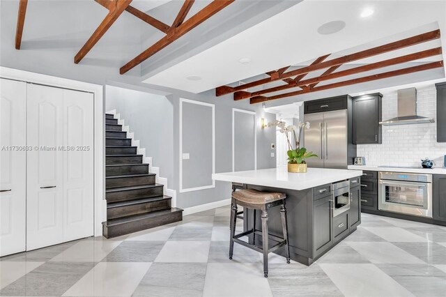 kitchen featuring a kitchen breakfast bar, a center island, built in appliances, beam ceiling, and wall chimney exhaust hood