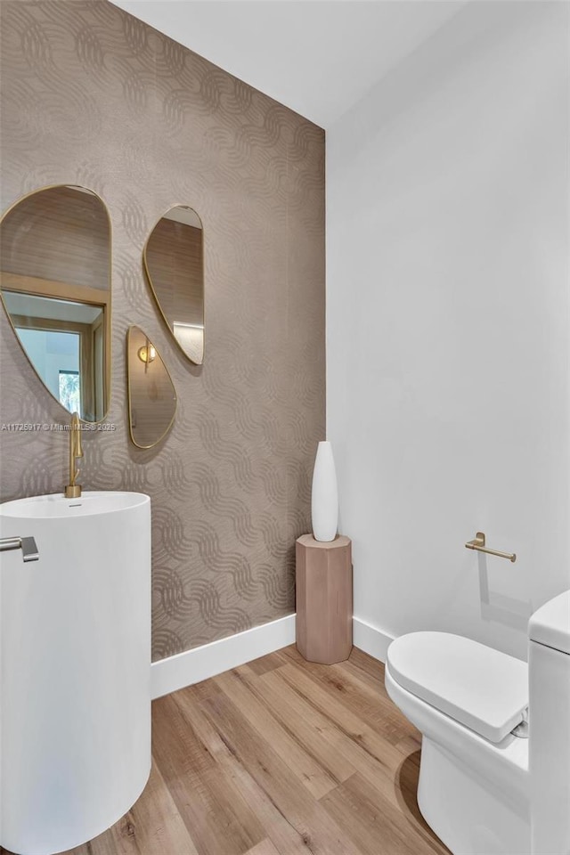 half bathroom featuring wood finished floors, toilet, baseboards, and wallpapered walls