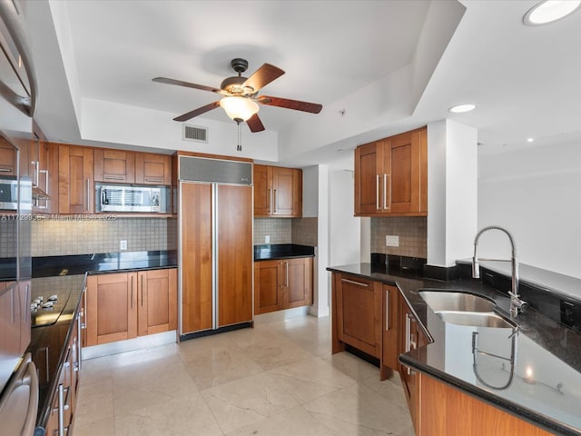 kitchen featuring sink, backsplash, built in appliances, a tray ceiling, and dark stone counters