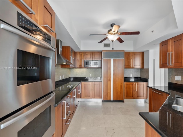 kitchen featuring a tray ceiling, decorative backsplash, stainless steel appliances, and wall chimney exhaust hood