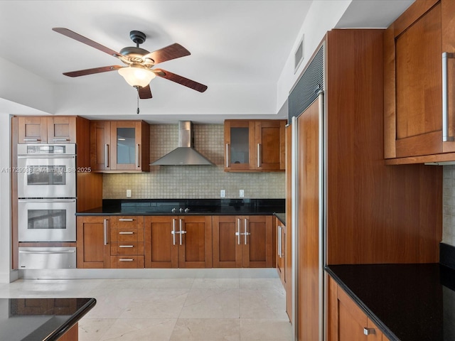 kitchen featuring wall chimney range hood, ceiling fan, double oven, backsplash, and paneled fridge