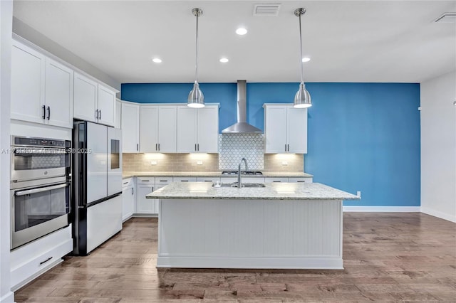 kitchen featuring wall chimney range hood, fridge, an island with sink, and white cabinets