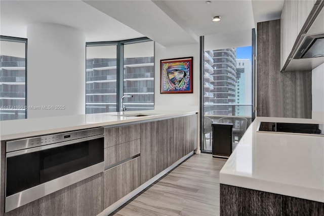 kitchen with sink, light wood-type flooring, a wall of windows, wall oven, and wall chimney range hood