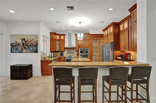 kitchen with appliances with stainless steel finishes, a kitchen bar, hanging light fixtures, kitchen peninsula, and wall chimney range hood