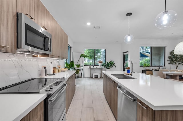 kitchen with sink, a kitchen island with sink, backsplash, stainless steel appliances, and decorative light fixtures