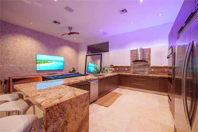 kitchen featuring white cabinetry, kitchen peninsula, stainless steel appliances, light stone countertops, and wall chimney exhaust hood