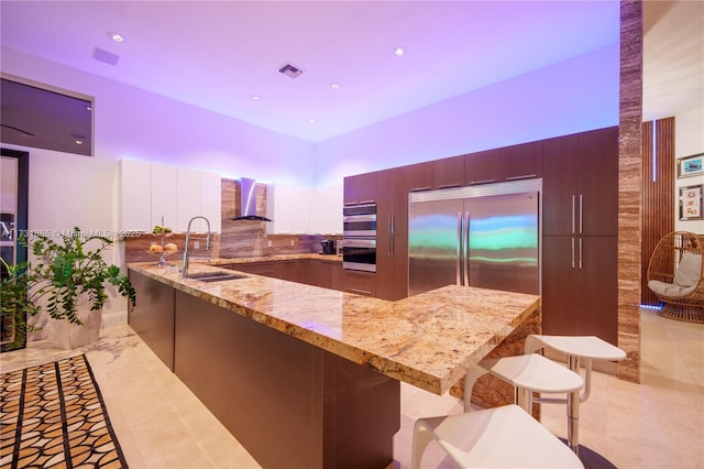 kitchen with wall chimney range hood, a breakfast bar area, light stone countertops, and built in refrigerator